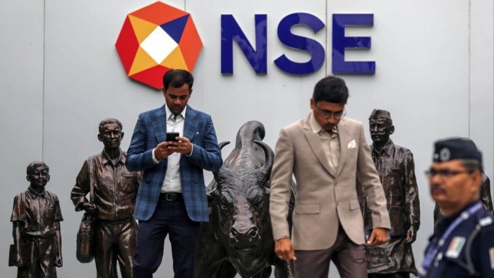 India on track for record IPO year A bronze statue of a bull is prominently displayed outside the National Stock Exchange in Mumbai. Two men in suits stand near the statue, one checking his phone, while a security officer is visible in the foreground. The NSE logo is mounted on the wall behind them.