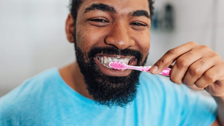 A person with black curly hair, a black bear and a bright blue tee brushing their teeth with a pink toothbrush.