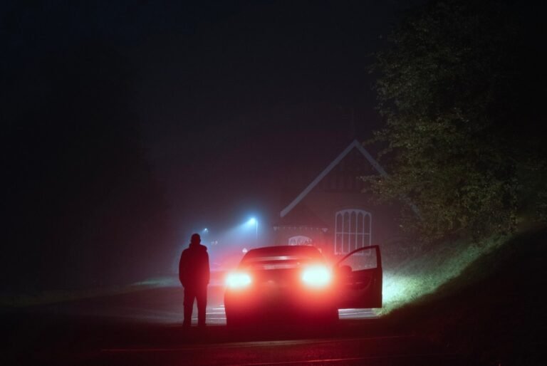 A mysterious person standing next to a car on a spooky empty road on a foggy night. Silhouetted by street lights.